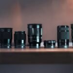 Lineup of Sony E-mount camera lenses arranged on a wooden table, including SIGMA 18-50mm F2.8, FE 35mm F1.4, and E 18-105mm F4 G